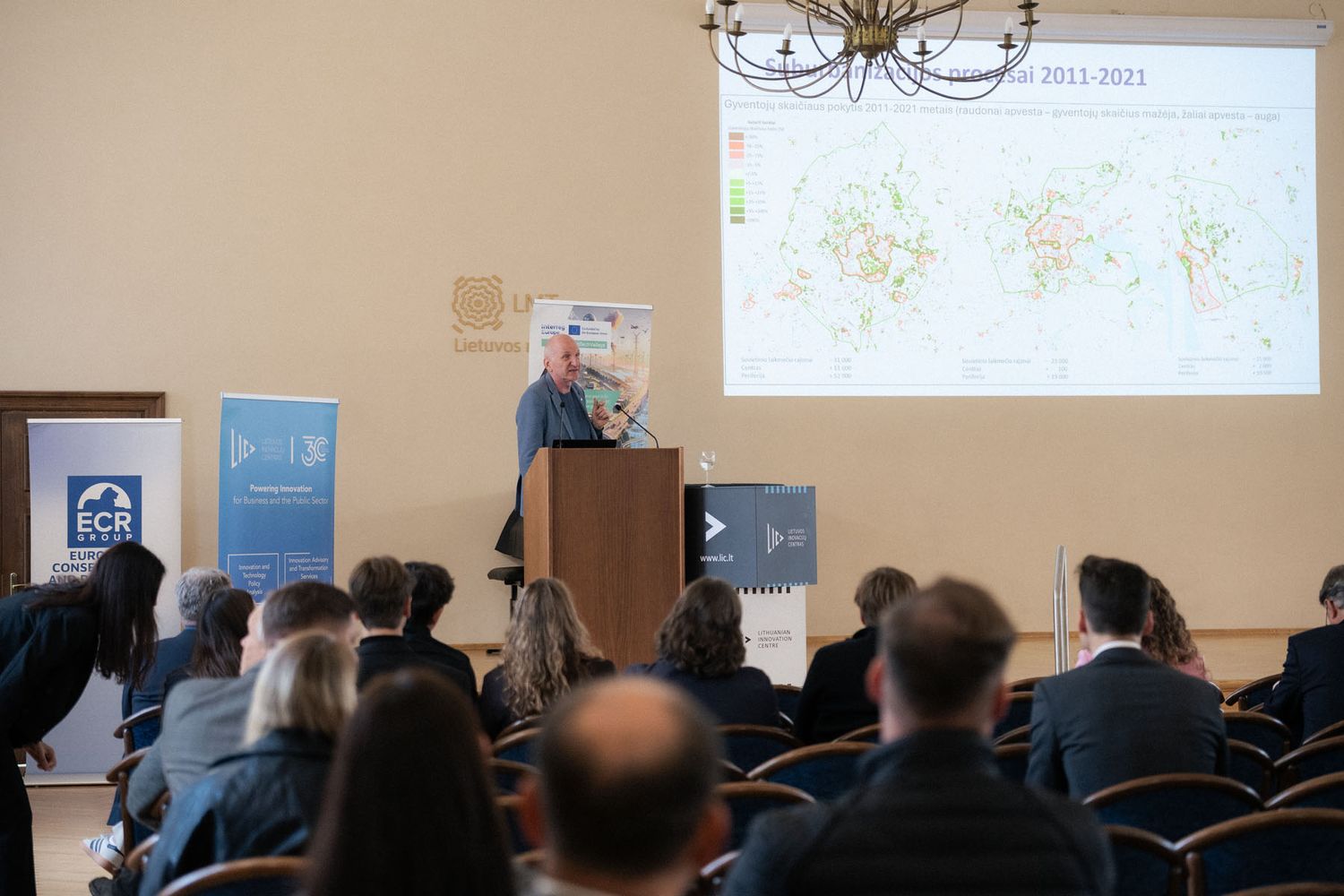Audience members watch a speaker at a lectern presenting a map visualization on a large screen in a historic conference hall.