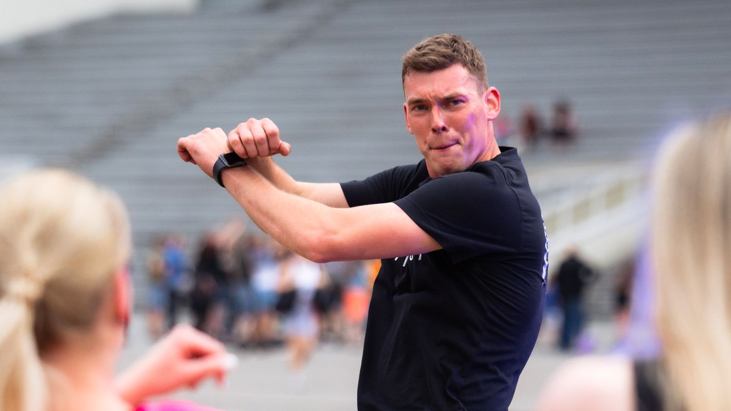 Man in a black t-shirt gestures with crossed arms while leading an outdoor fitness class near a stadium.