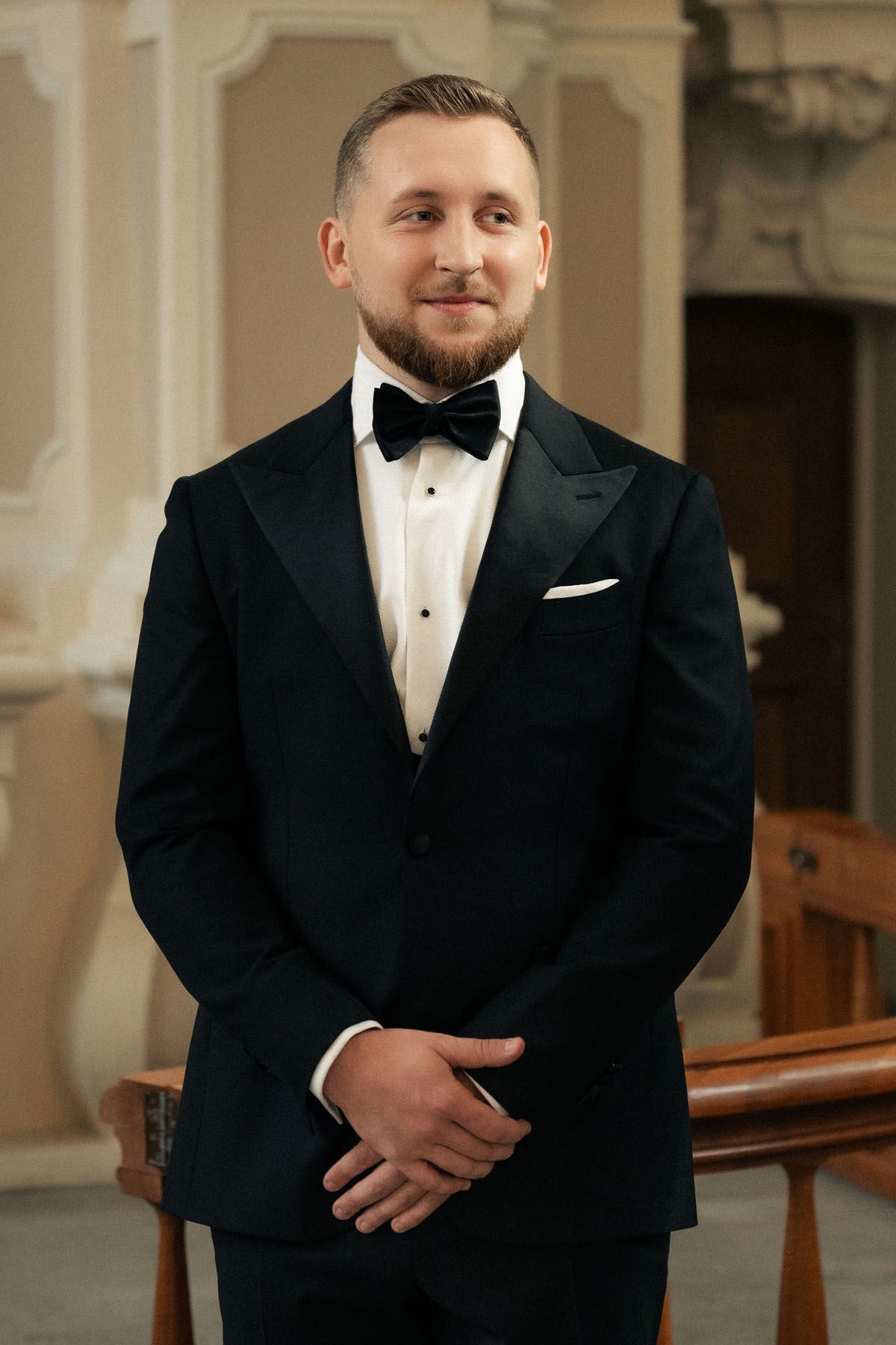 Smiling groom with a beard wears a black tuxedo with bow tie and white pocket square while waiting in a church.