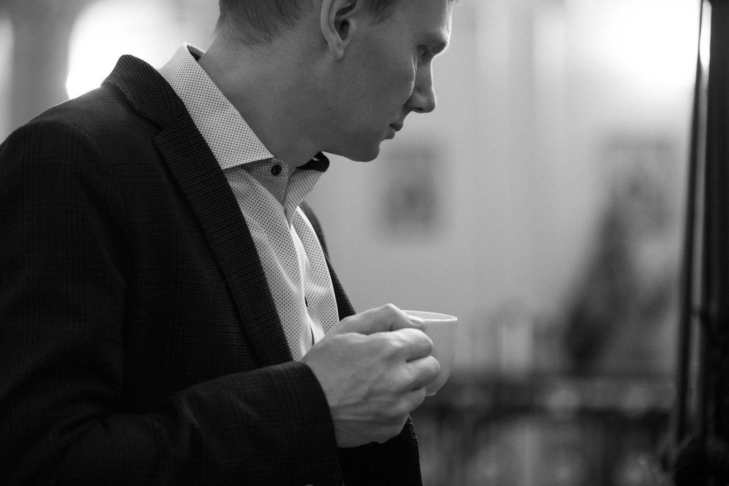 Black and white profile of a man in a dark blazer and open-collar shirt looking down at a small cup he holds.