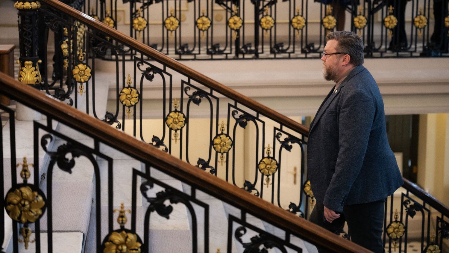 A bearded man in a dark blazer walks up a staircase with ornate black and gold railings.