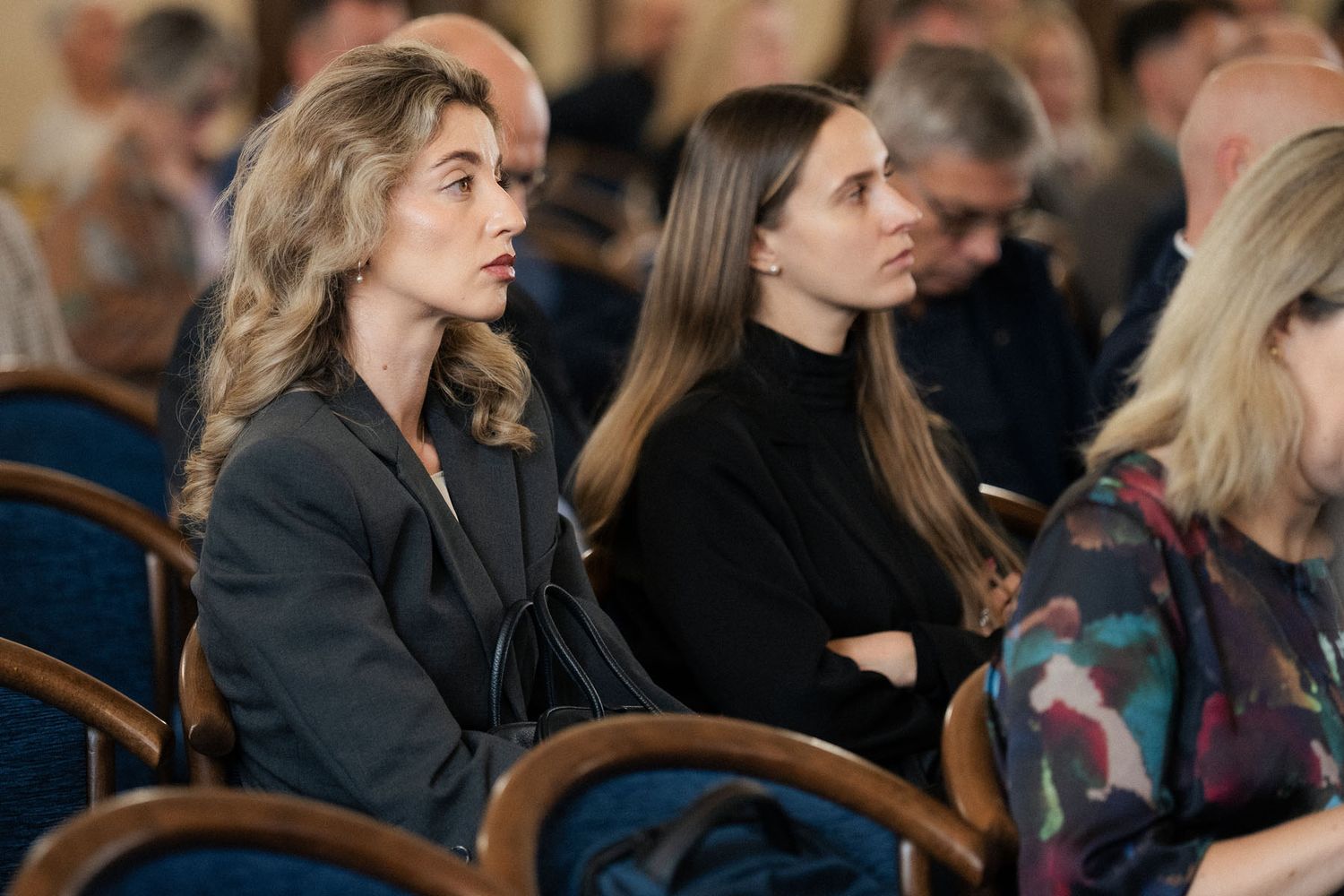 Two attentive women with long hair sit among conference attendees in a hall, listening to a presentation.
