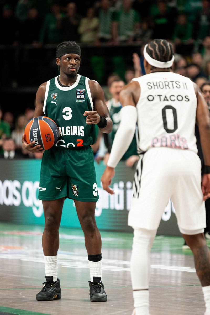 Basketball player in a green number 3 jersey holds the ball while facing a defender in white number 0 during a game in an arena.