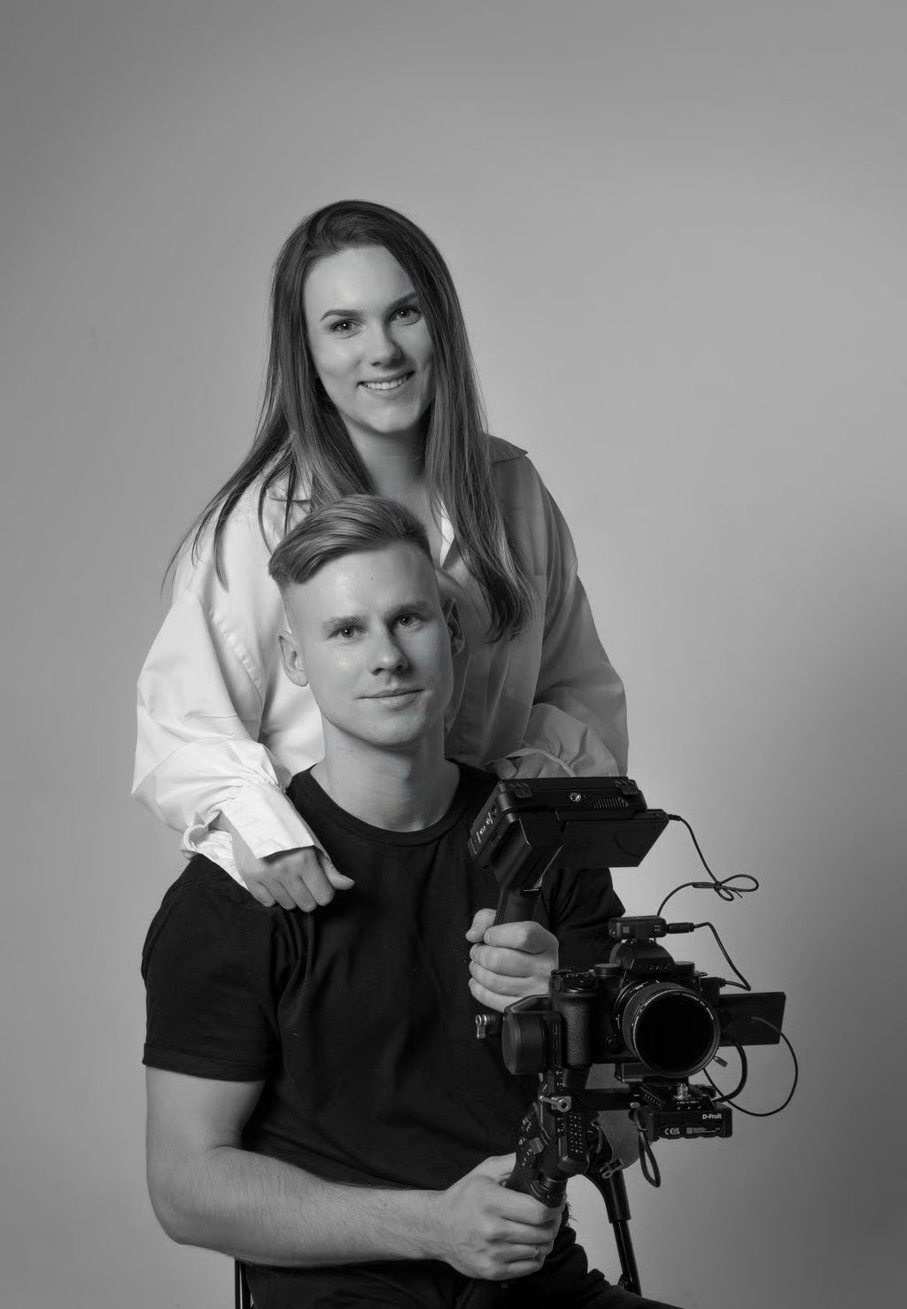 A black and white studio portrait of a smiling woman standing behind a man holding a professional video camera.
