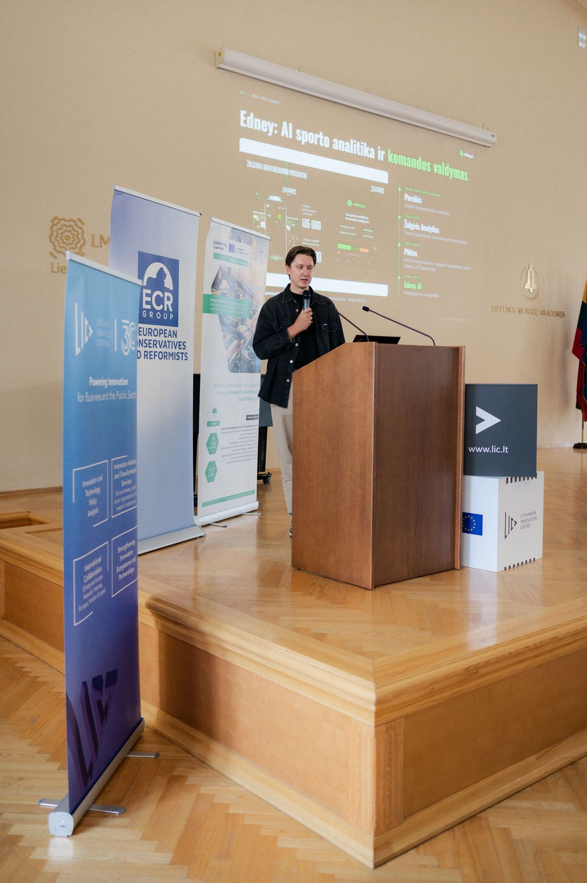 Man speaking at a wooden podium beside event banners and a projected slide about AI sports analytics.