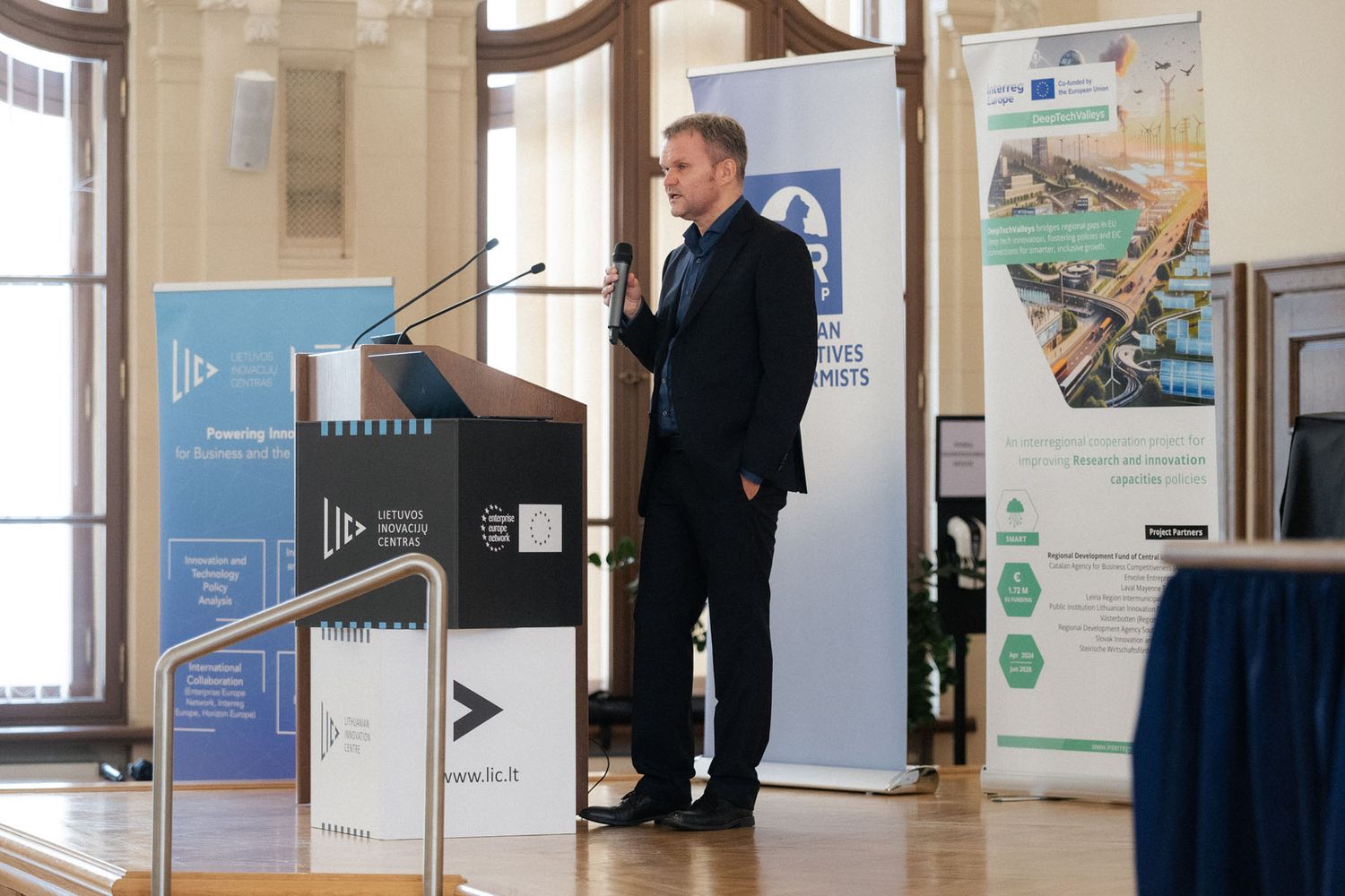 A man in a dark suit stands on a stage holding a microphone beside a podium and event banners in a sunlit hall.