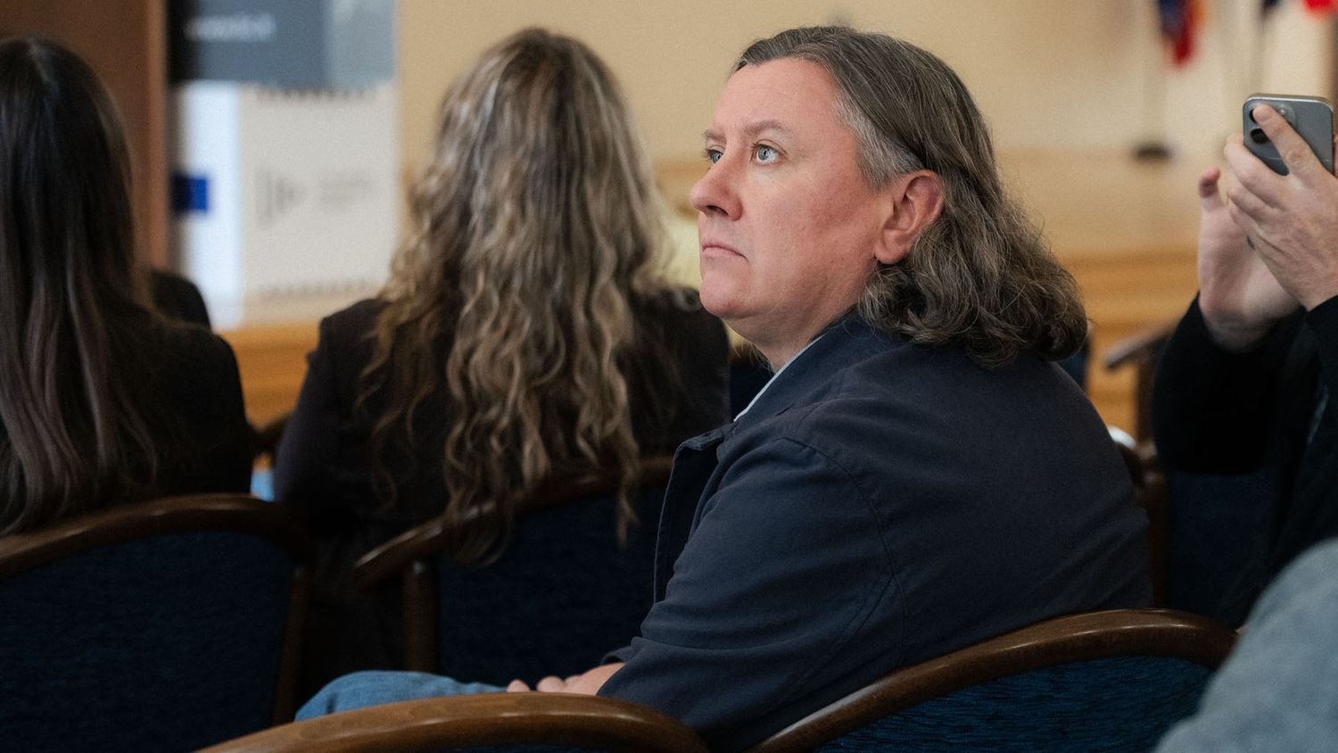 Woman with long gray hair sits in a blue chair listening attentively among attendees at a conference event.