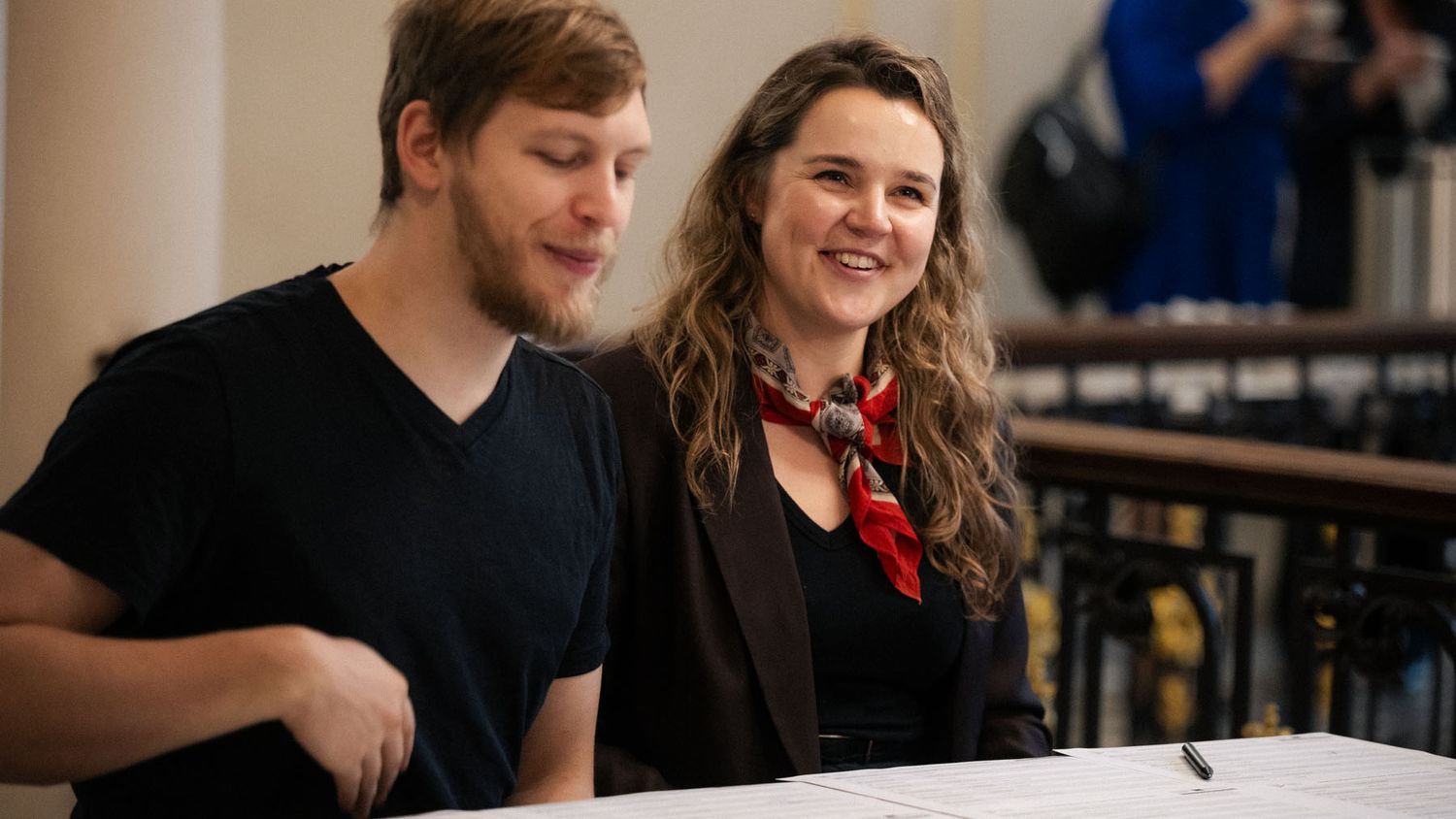 A smiling woman in a red scarf sits beside a bearded man in a black t-shirt at a table during an indoor event.