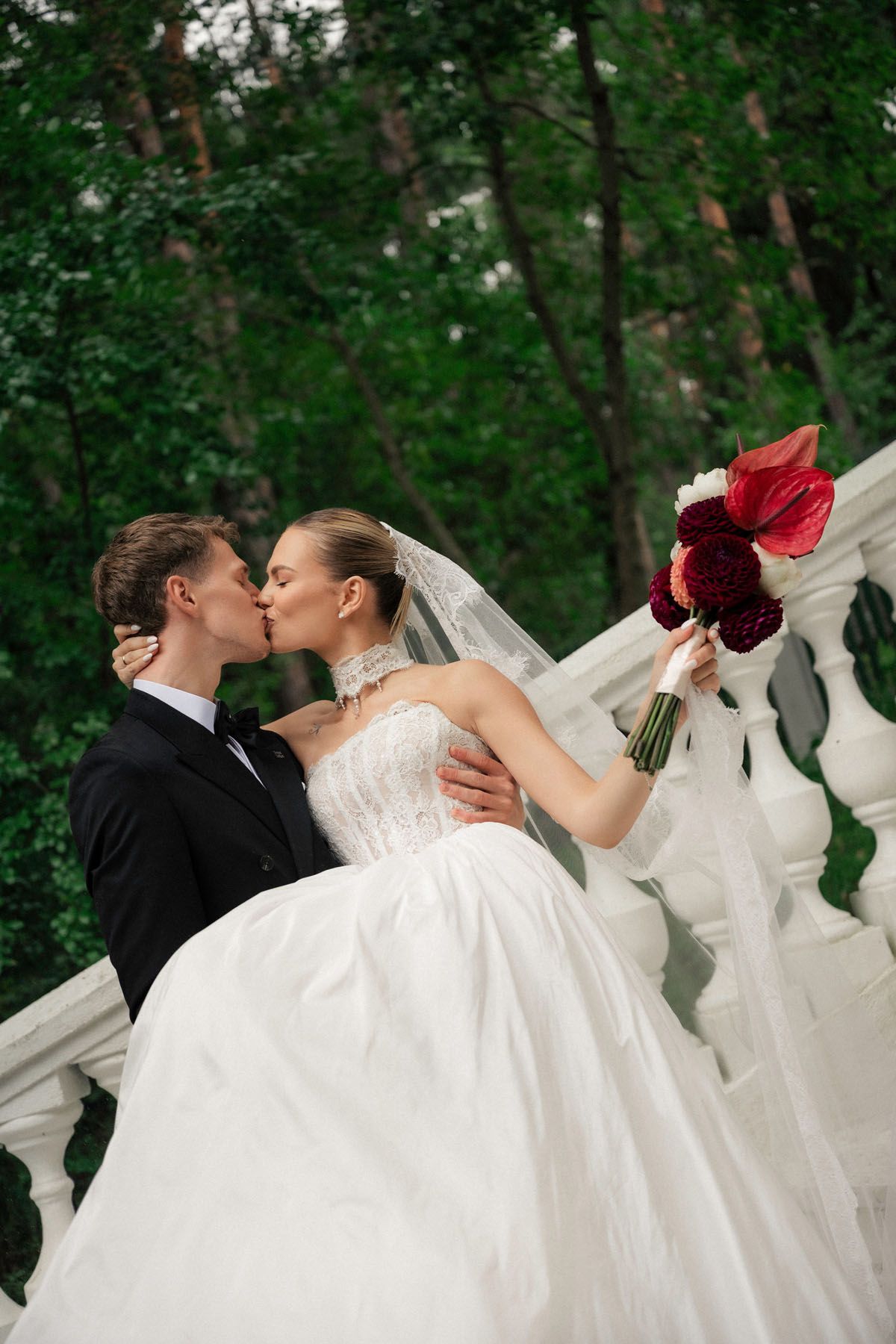 Bride in a white gown and groom in a black suit kiss on a stone staircase among green trees, holding a red bouquet.
