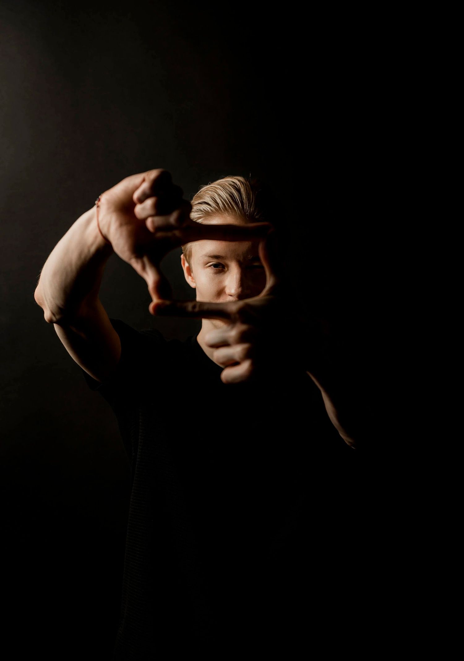 Young man with blond hair forms a camera frame with his hands against a dark background in a moody studio portrait.