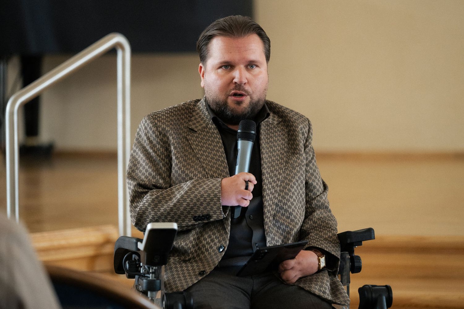A man in a tweed blazer sits in a wheelchair and speaks into a microphone during a conference discussion.