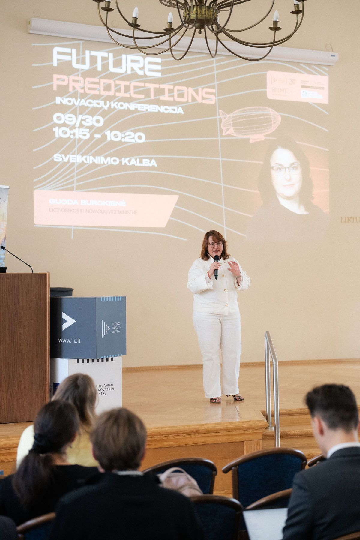 Woman in white suit speaks into a microphone on stage beside a screen showing Future Predictions while attendees watch.