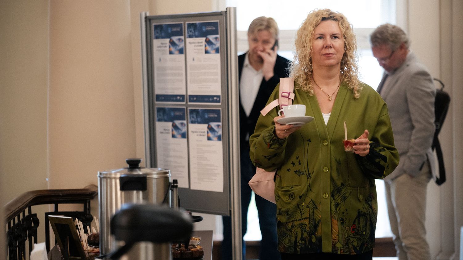 A woman in a green jacket holds a cup and saucer at a coffee station while attendees chat behind her.