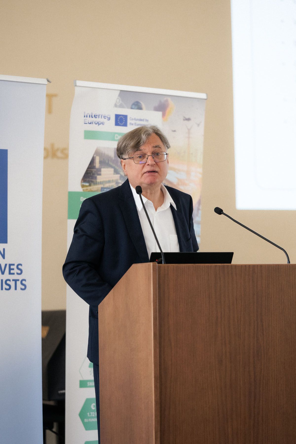 Man with glasses and a dark blazer speaks at a wooden podium with microphones in front of Interreg Europe and ECR banners.