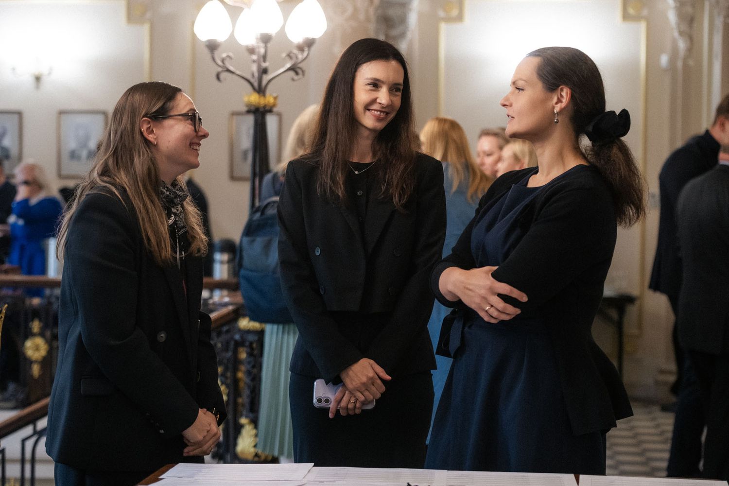 Three women in dark clothing chat and smile at a conference networking event in an elegant, chandelier-lit hall.