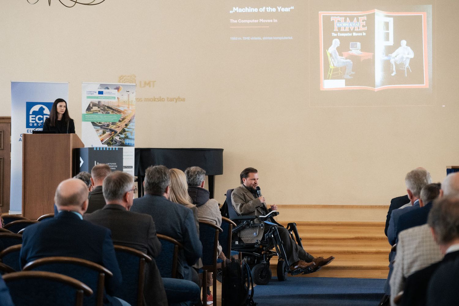 Attendees watch a man in a wheelchair speaking while a woman stands at a podium and a presentation slide shows on the wall.