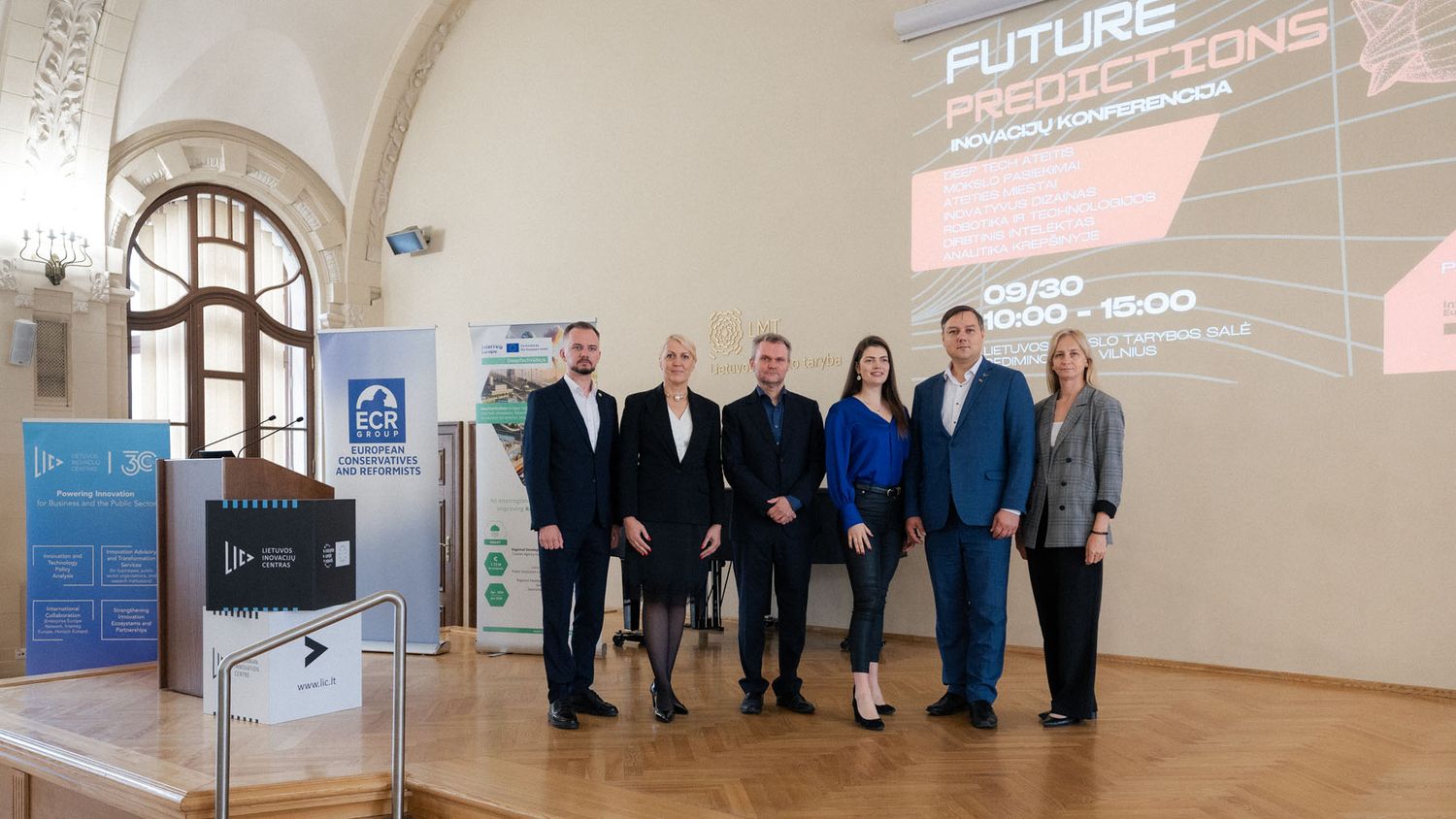Six smiling professionals stand together on stage in front of a Future Predictions conference screen.