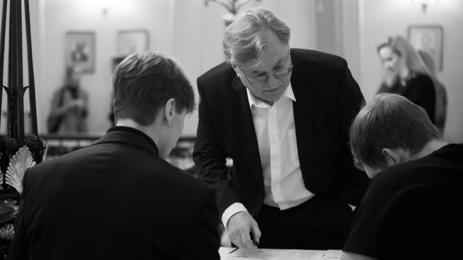 A black-and-white photo of a man in a dark suit leaning over a table to speak with two seated attendees.
