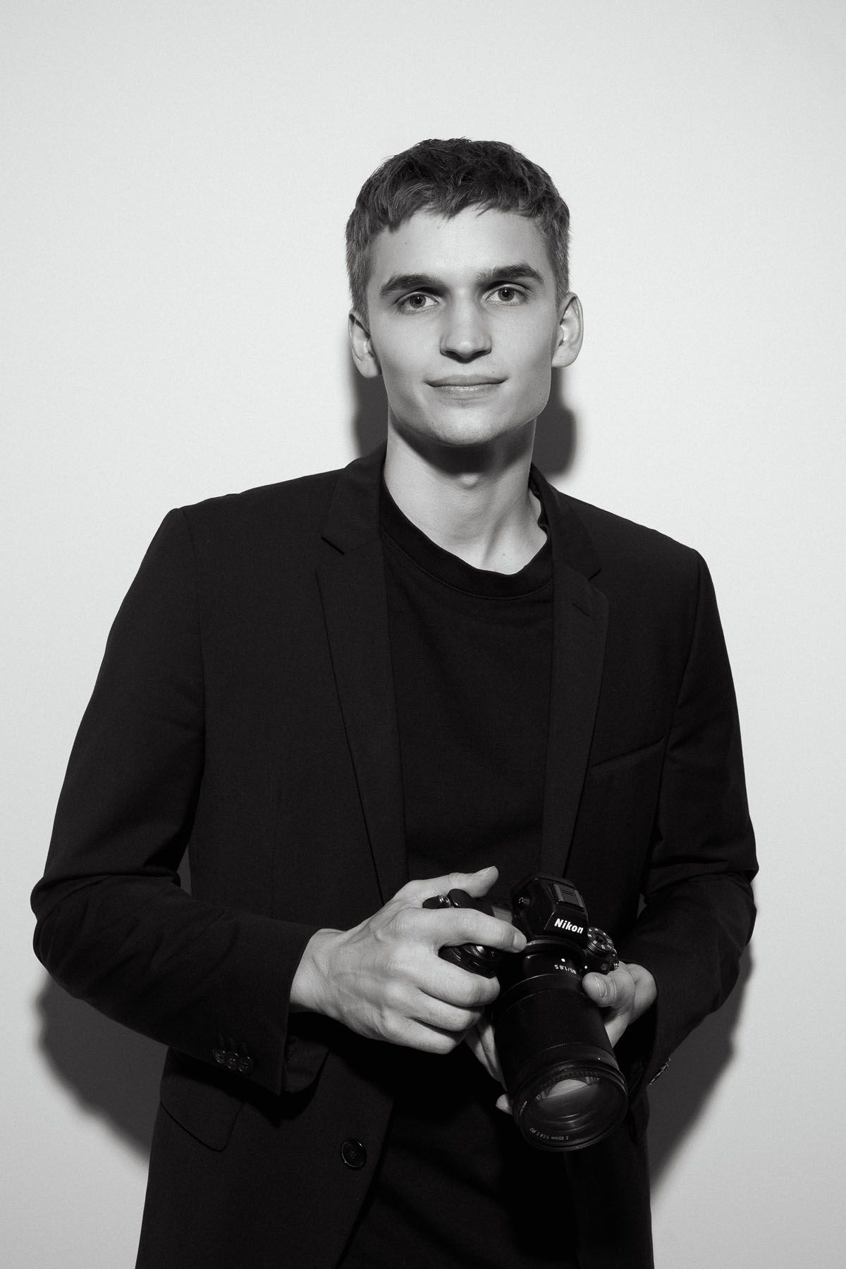 Monochrome portrait of a young photographer in a black jacket and tee holding a Nikon camera in a studio.