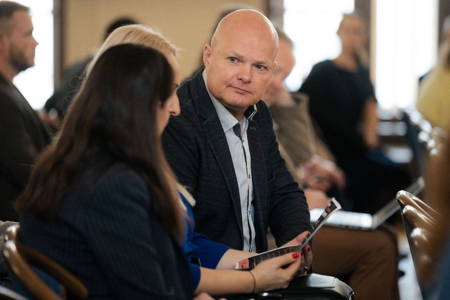 A bald man in a dark blazer sits among attendees reading a document while a woman beside him looks on at a conference.