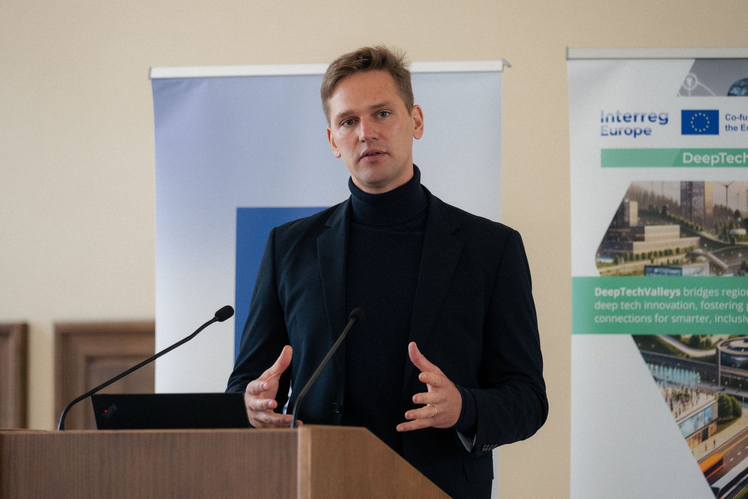 A man in a black turtleneck and blazer gestures while speaking at a conference podium with a DeepTechBridge banner behind him.