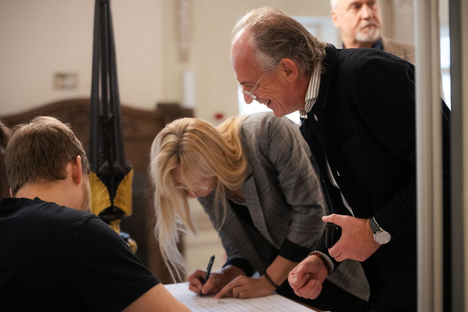 A blonde woman signs a registration form as a man in a dark jacket leans in to watch beside her.