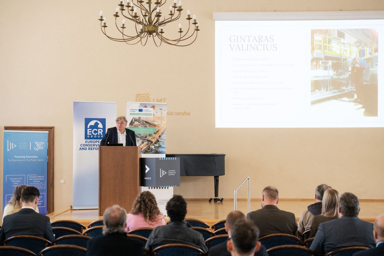Audience seated in a conference hall listens to a speaker at a podium beside a presentation slide and ECR Group banner.