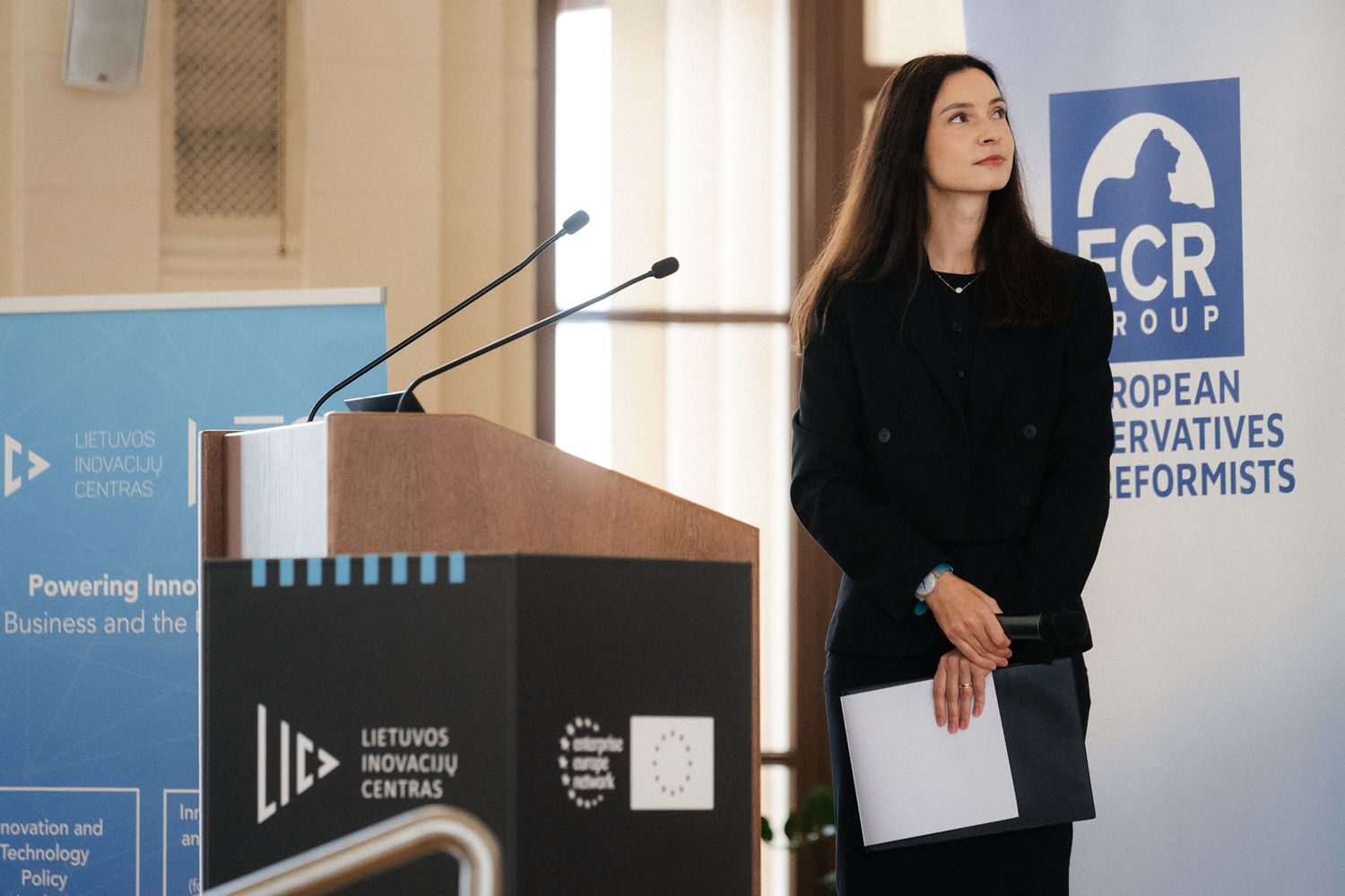 Woman in a dark blazer stands beside a podium holding a folder at an ECR Group conference on innovation in Lithuania.