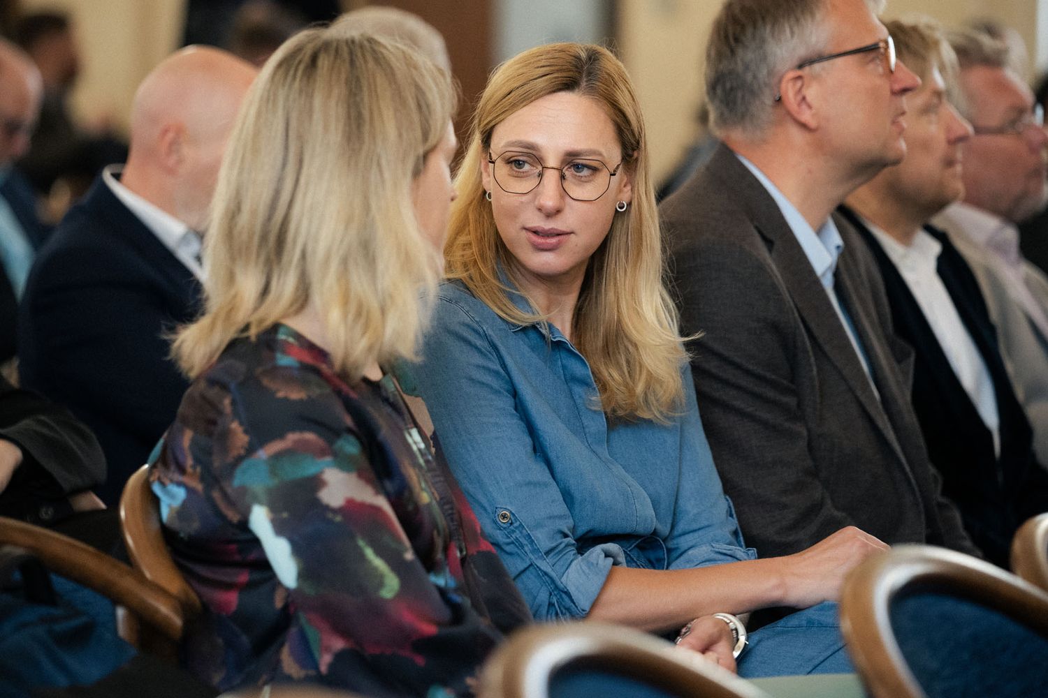 Two women in conversation sit in a conference audience, one with glasses and long blonde hair, with other attendees nearby.