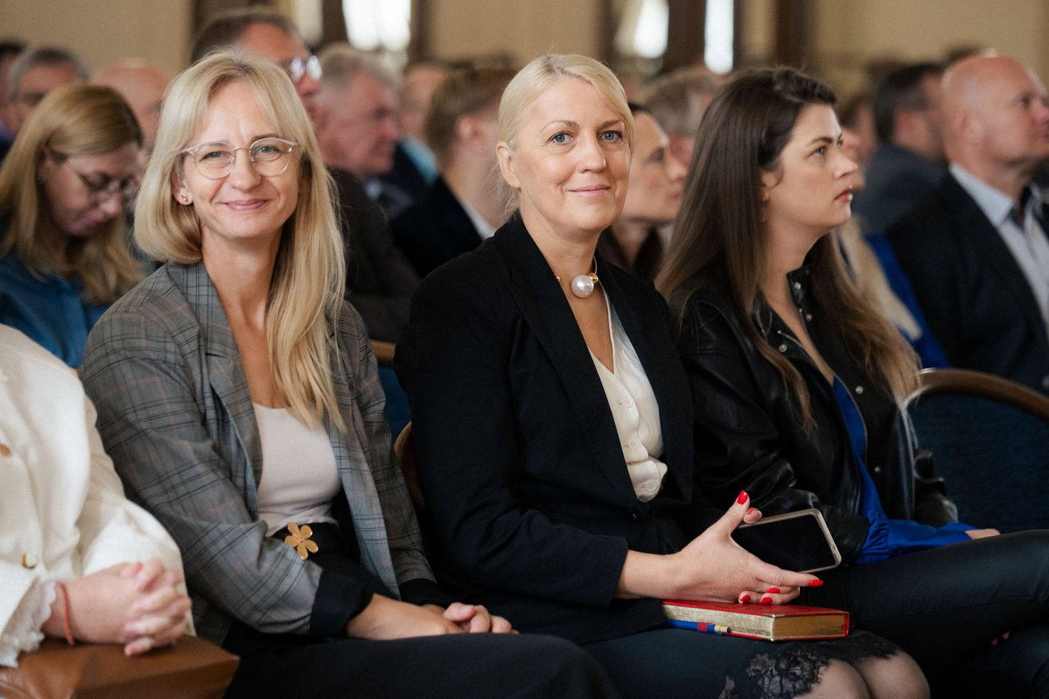 Smiling women in blazers sit among attendees in a conference hall, one holding a smartphone, during an event.