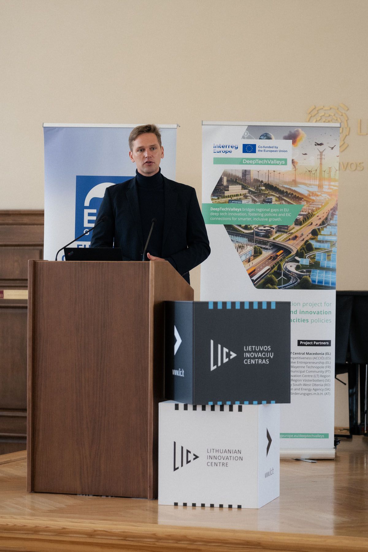 A speaker in a dark blazer stands at a wooden podium beside Lithuanian Innovation Centre boxes and an Interreg Europe banner.