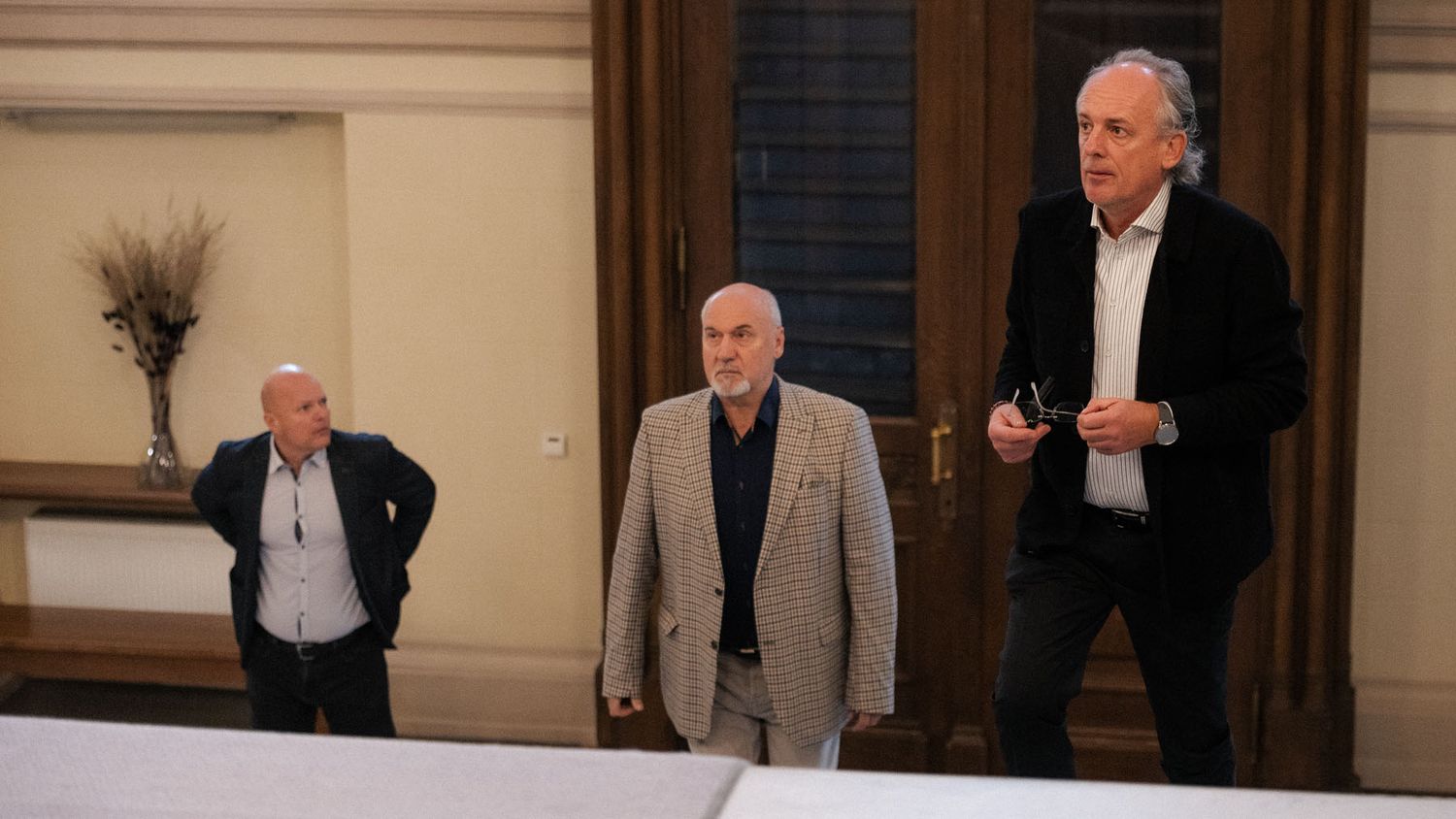 Three men in jackets stand in an elegant hall with wooden doors during a conference event.
