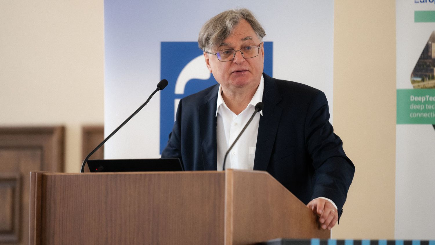 Close-up of a gray-haired man in glasses and a navy blazer speaking at a wooden podium with two microphones.