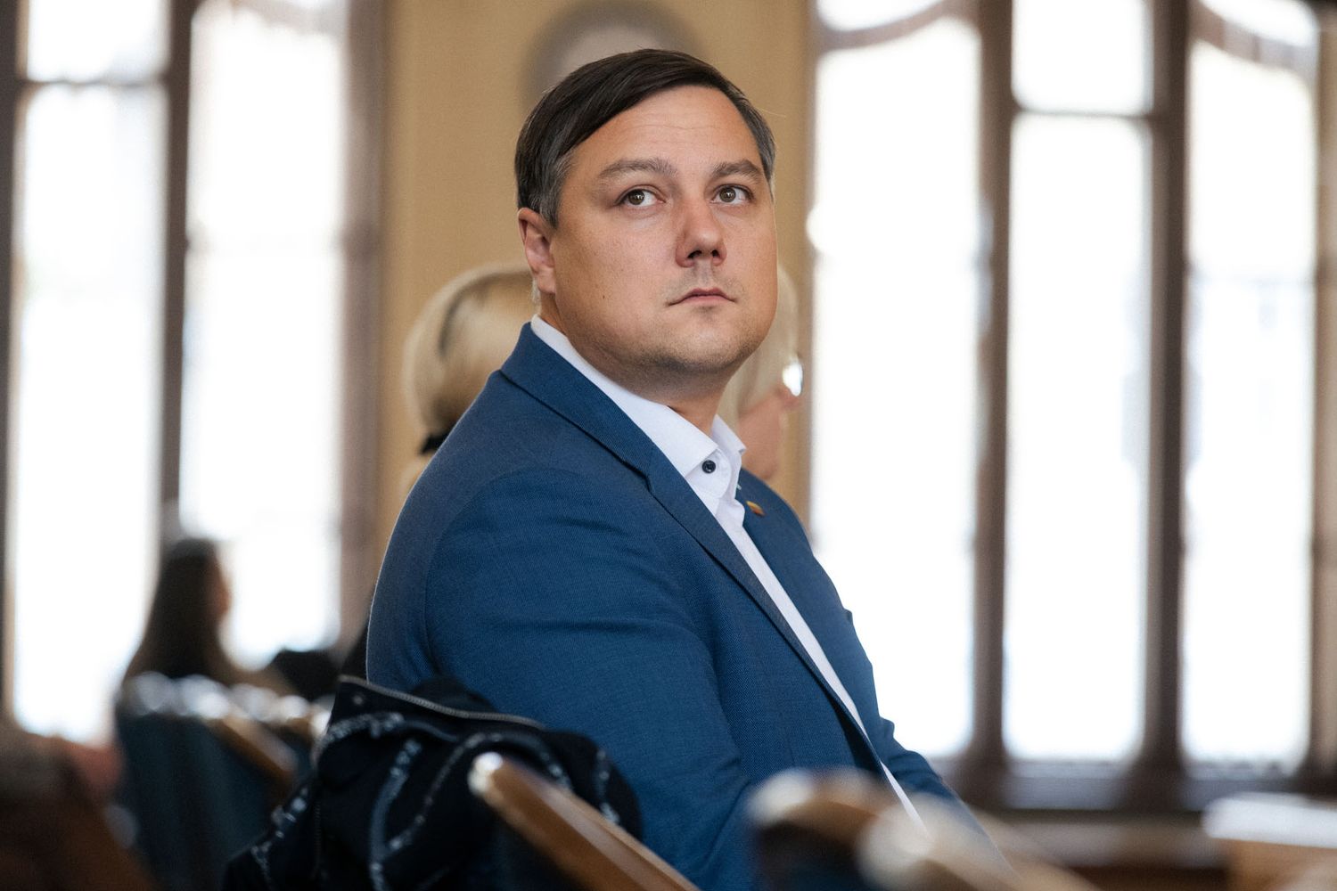 Man in a blue blazer sits thoughtfully in a conference audience inside a historic hall with tall windows.