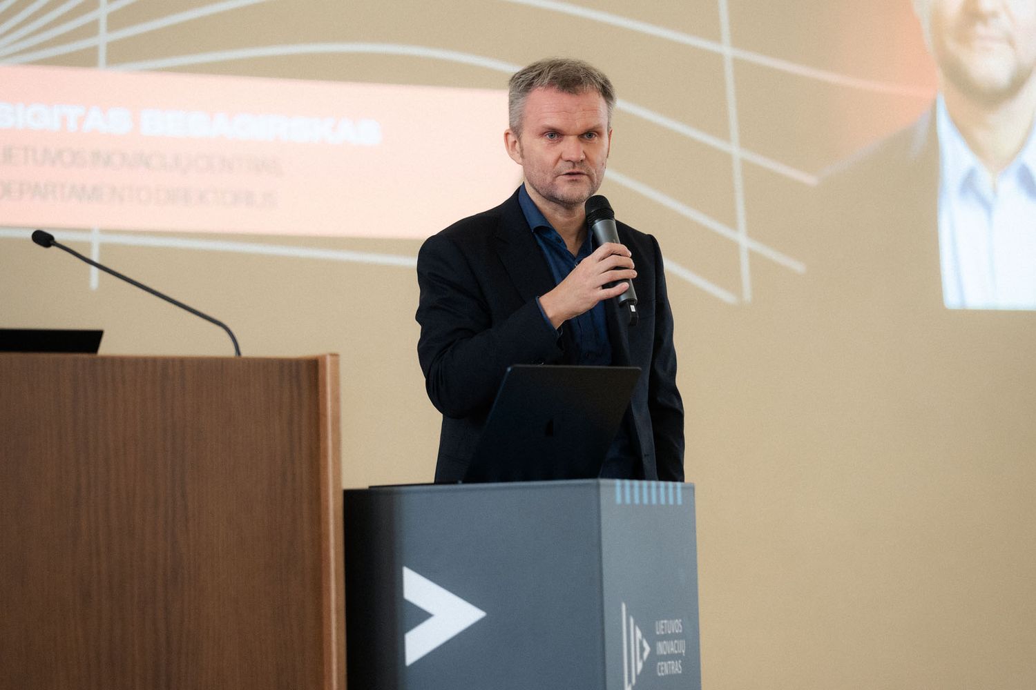 A man in a dark blazer speaks into a microphone beside a podium at a conference with a presentation screen behind him.