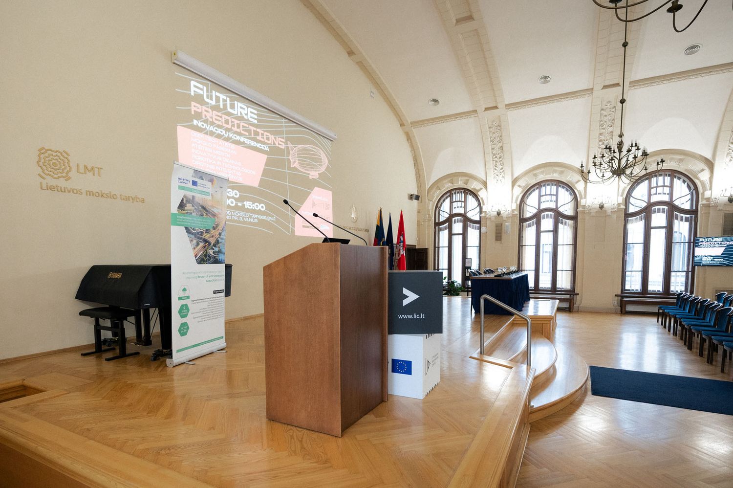 Wooden podium with microphones on a stage beside a piano, banners and flags in a grand hall with arched windows.