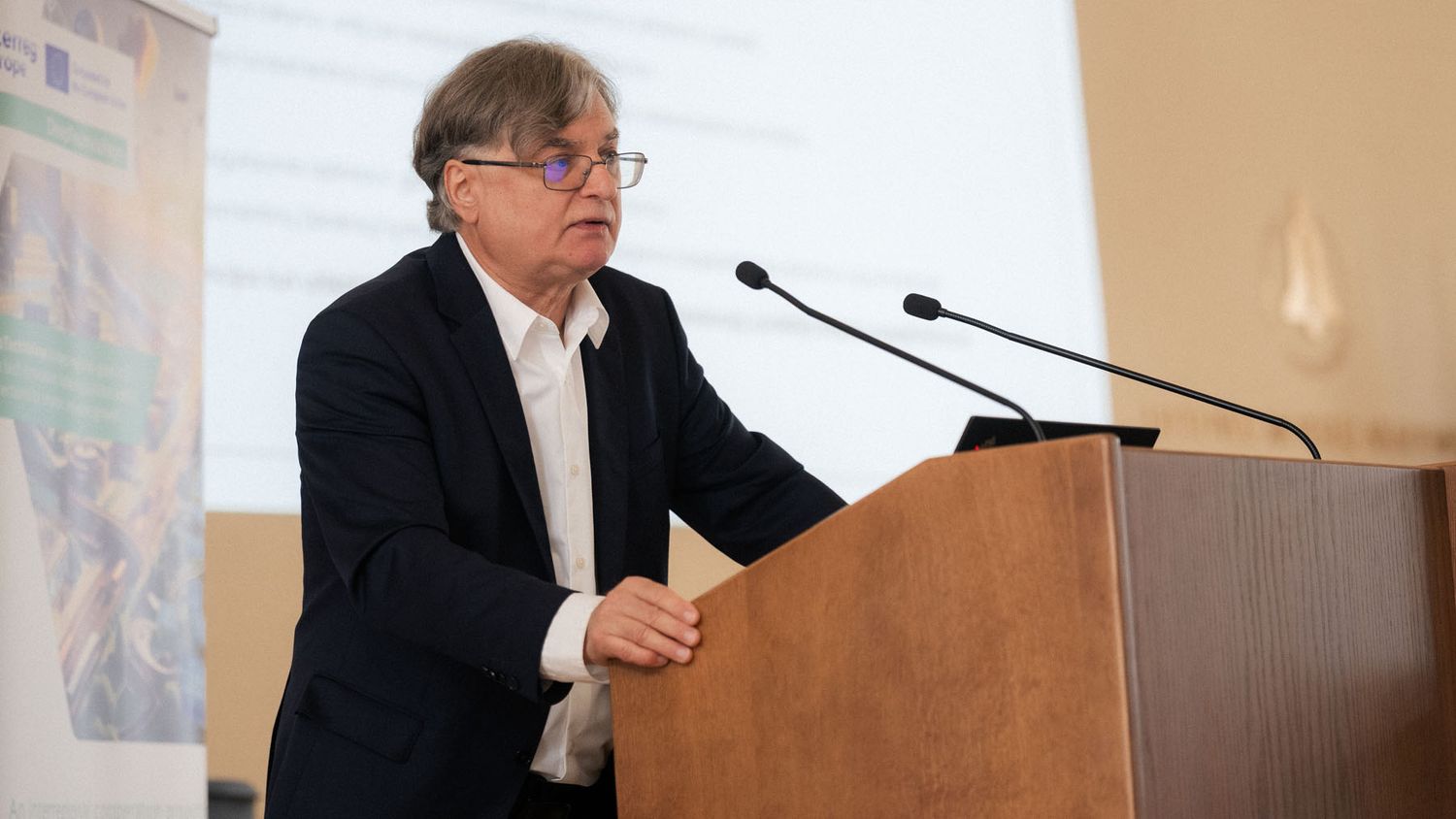 Man in a dark blazer and glasses speaks at a wooden podium with two microphones during a conference presentation.