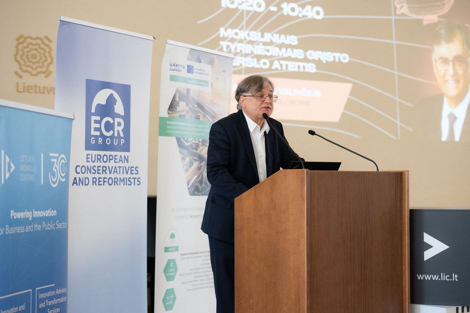 Speaker in a dark jacket stands at a podium before ECR Group European Conservatives and Reformists banners at a conference.