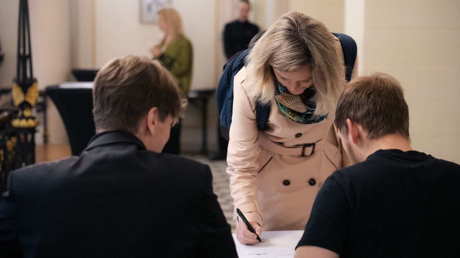 A woman in a beige trench coat signs a registration sheet at a table while two men stand beside her.