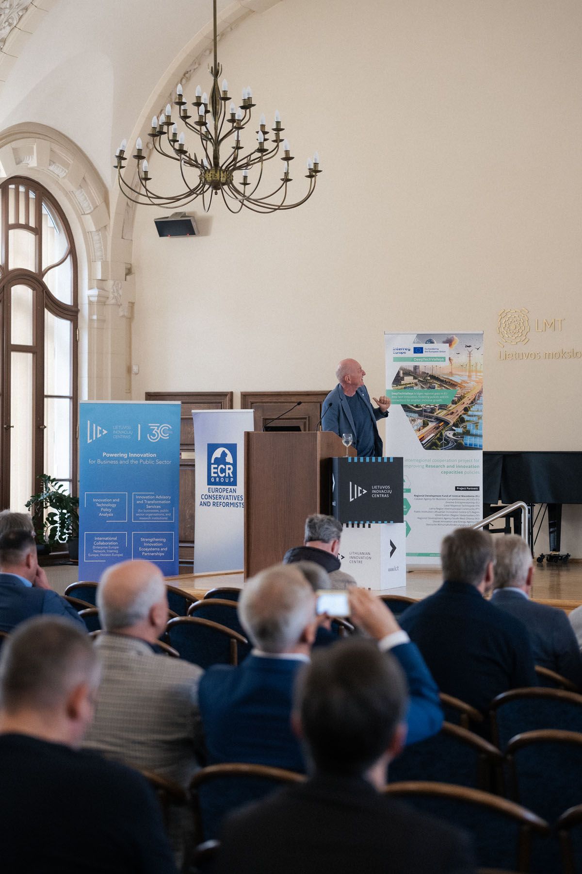 A speaker stands at a lectern in an ornate hall with a chandelier, facing seated attendees at an innovation conference.