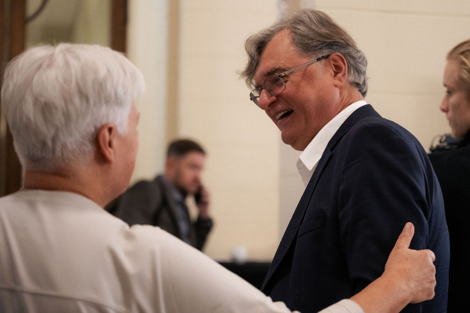 A smiling man in glasses and a navy blazer shares a warm moment with a gray-haired woman who rests a hand on his shoulder.