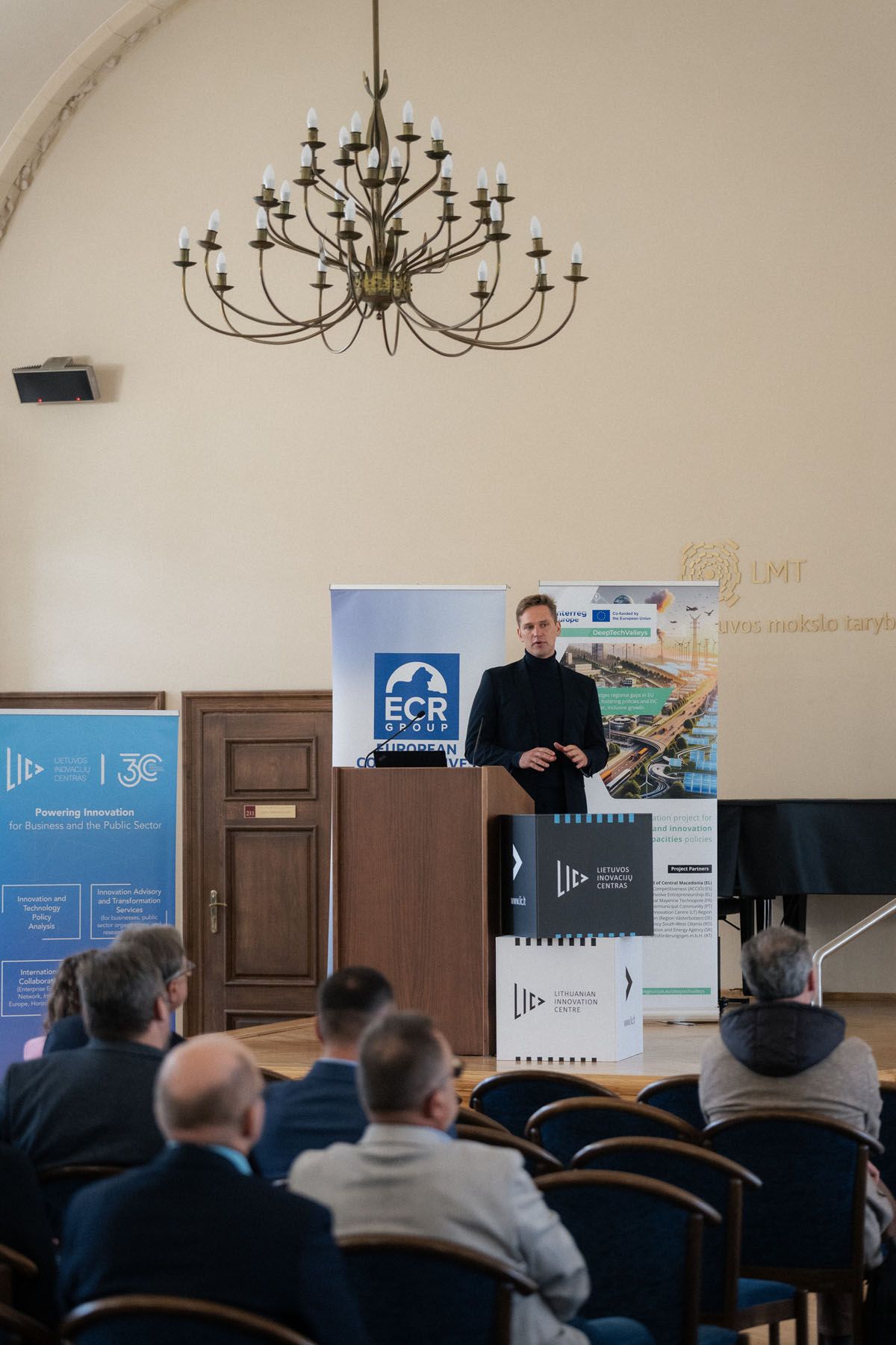 Speaker in dark clothing stands at a wooden podium beneath a chandelier while seated attendees watch at a conference.