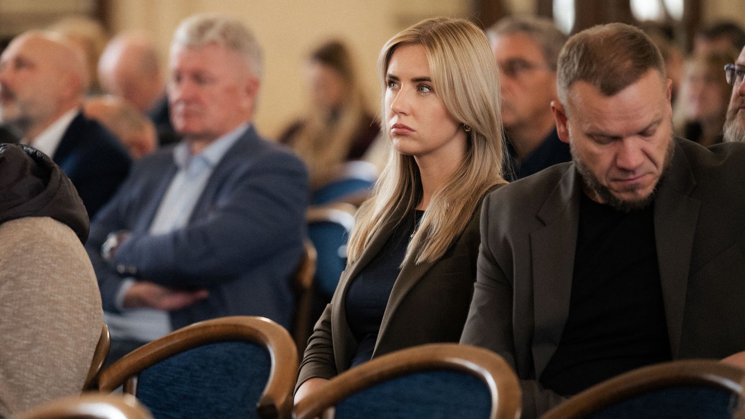 Attendees sit in blue wooden chairs at a conference, with a blonde woman and a man in a dark jacket looking ahead.