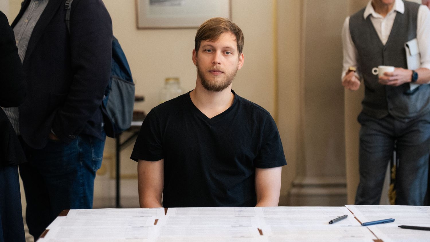 A bearded man in a black t-shirt sits at a registration table with papers and pens at an indoor event.