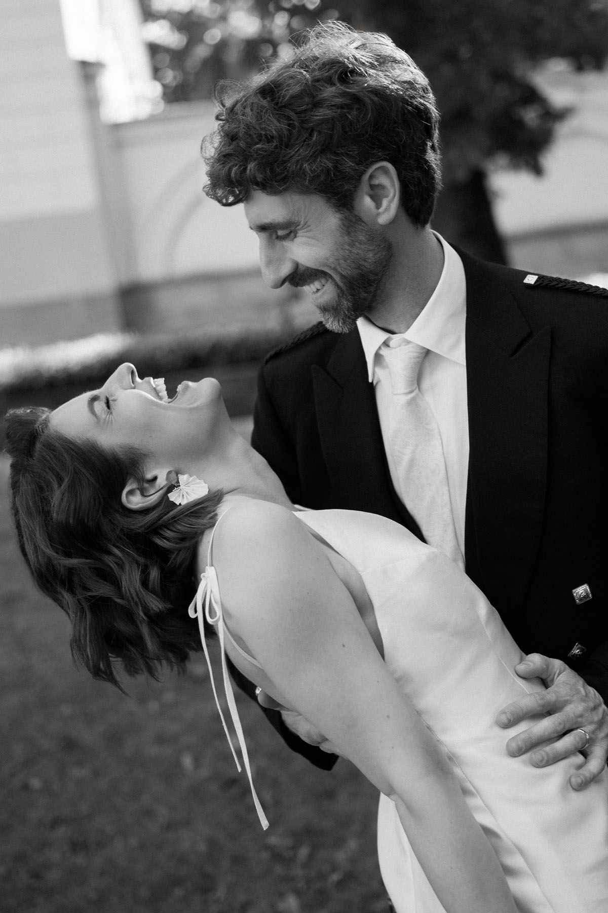 Bride laughing joyfully with her head tilted back as the groom in a dark jacket smiles and holds her in an outdoor wedding portrait.