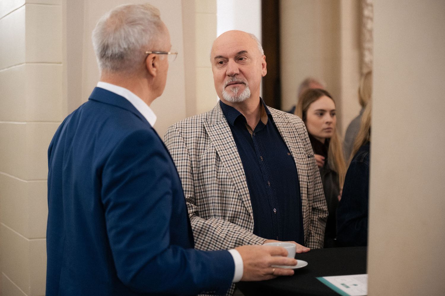 A man in a blue suit talks with a bearded man in a checked blazer as other attendees stand behind them at an event.
