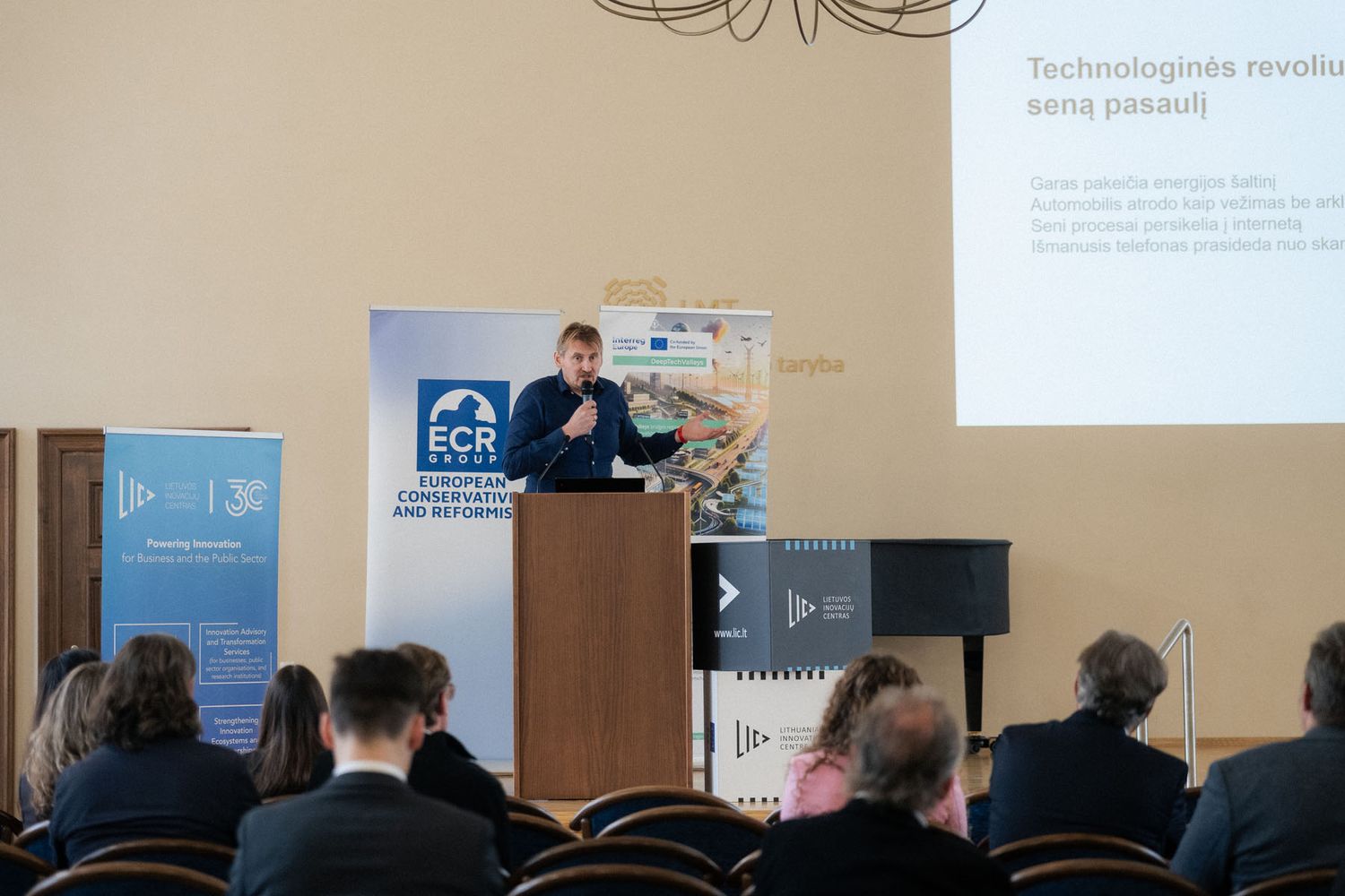 A presenter speaks at a podium in front of an audience, with a slide on technology revolution projected on the wall.