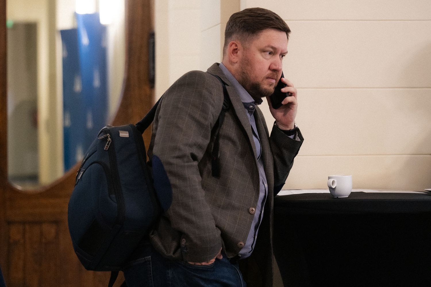 A bearded man in a gray jacket carrying a navy backpack talks on his phone near a table with a white coffee cup.