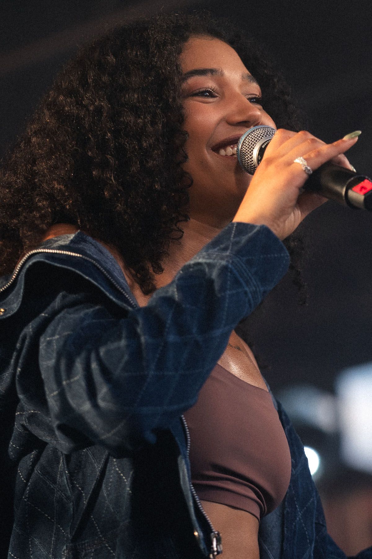 Smiling woman with curly hair sings into a microphone, wearing a quilted denim jacket during a live performance.