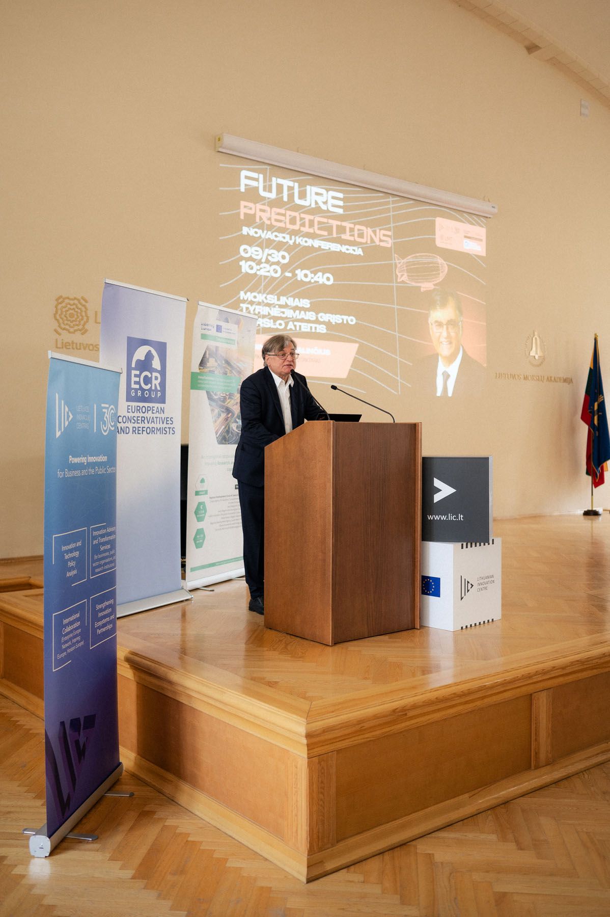 Man in a dark suit speaks at a wooden podium beside event banners, with a Future Predictions slide on the screen.